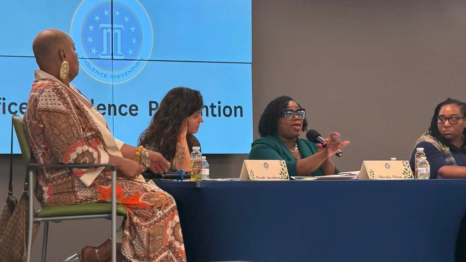 Nikesha Tilton speaking into a microphone from a panel table at an Office of Violence Prevention event, seated with three fellow panelists