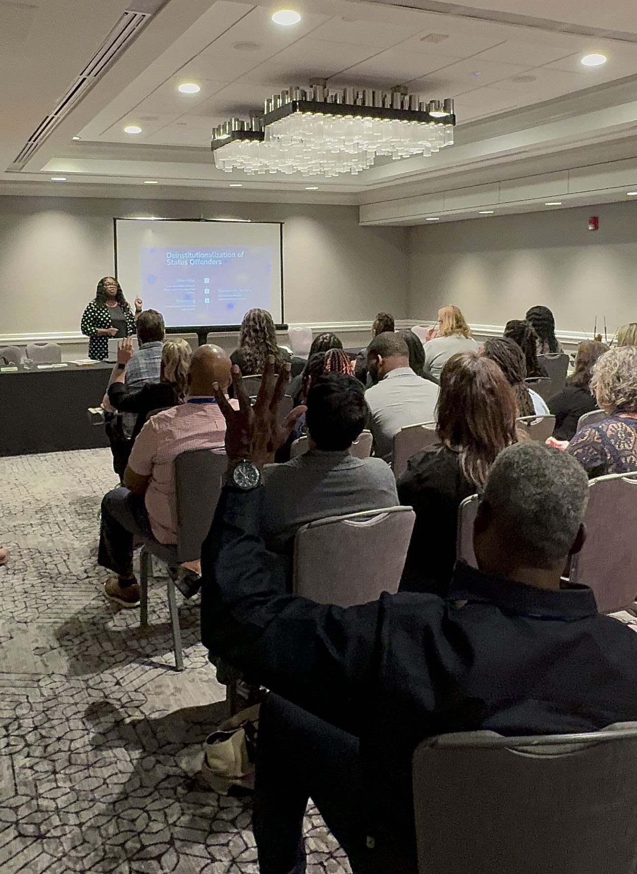 Nikesha Tilton presenting at the front of a conference room while participants listen and raise their hands